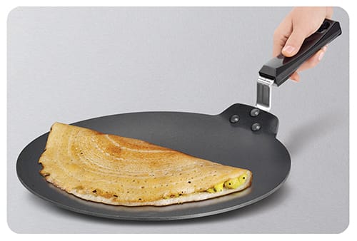A folded, golden dosa cooking on a nonstick tava griddle held by its handle