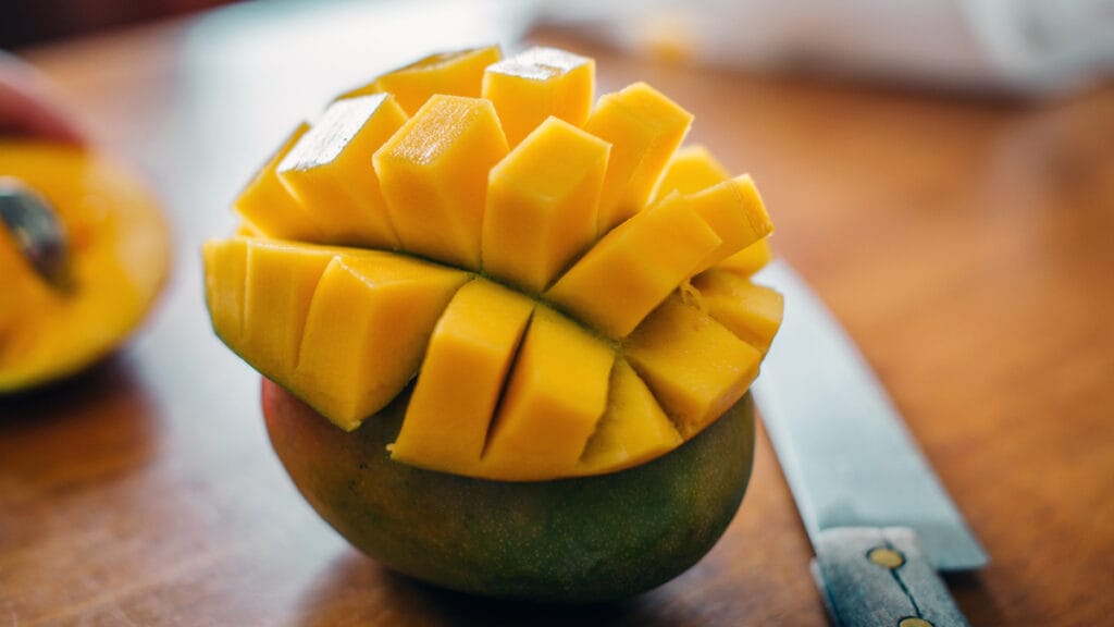 A ripe mango scored into cubes and pushed inside-out, with a paring knife on a wooden board