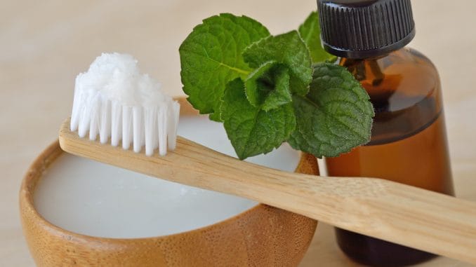 Homemade peppermint toothpaste on a bamboo toothbrush beside fresh mint leaves and an amber essential-oil bottle