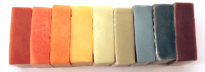 Row of eight handmade soap bars tinted with natural colorants in earthy reds, oranges, yellows, and blues