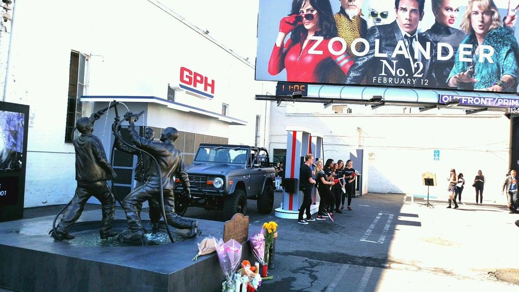 Outdoor bronze statue of gas-station attendants beside a Zoolander No. 2 movie billboard
